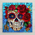 Stained glass style ceramic art tile of a Day of the Dead sugar skull decorated with red roses and gold accents on a blue leaded-glass background — a Dia de los Muertos decoration.