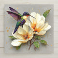 Ceramic art tile depicting a broadtail hummingbird hovering beside large creamy white magnolia blooms in fine art oil painting style with gold leaf accents, warm ivory and champagne gold tones