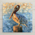 Sea glass style ceramic tile of a lifelike brown pelican perched on a weathered wooden piling with mosaic glass feathers against a blue and gold sea glass background