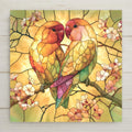Stained glass style ceramic tile of two lovebirds with rosy faces and gold and green feathers perched close together on a flowering branch in radiant golden light