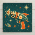 Retro atomic age illustration ceramic tile of a vintage toy ray gun with fins and rings firing a starburst zap among stars and a ringed planet in teal orange and cream