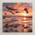 Stained glass style ceramic art tile of sandhill cranes flying over a marsh at sunset, orange and pink glass clouds above wading cranes and a golden sun path on the water.